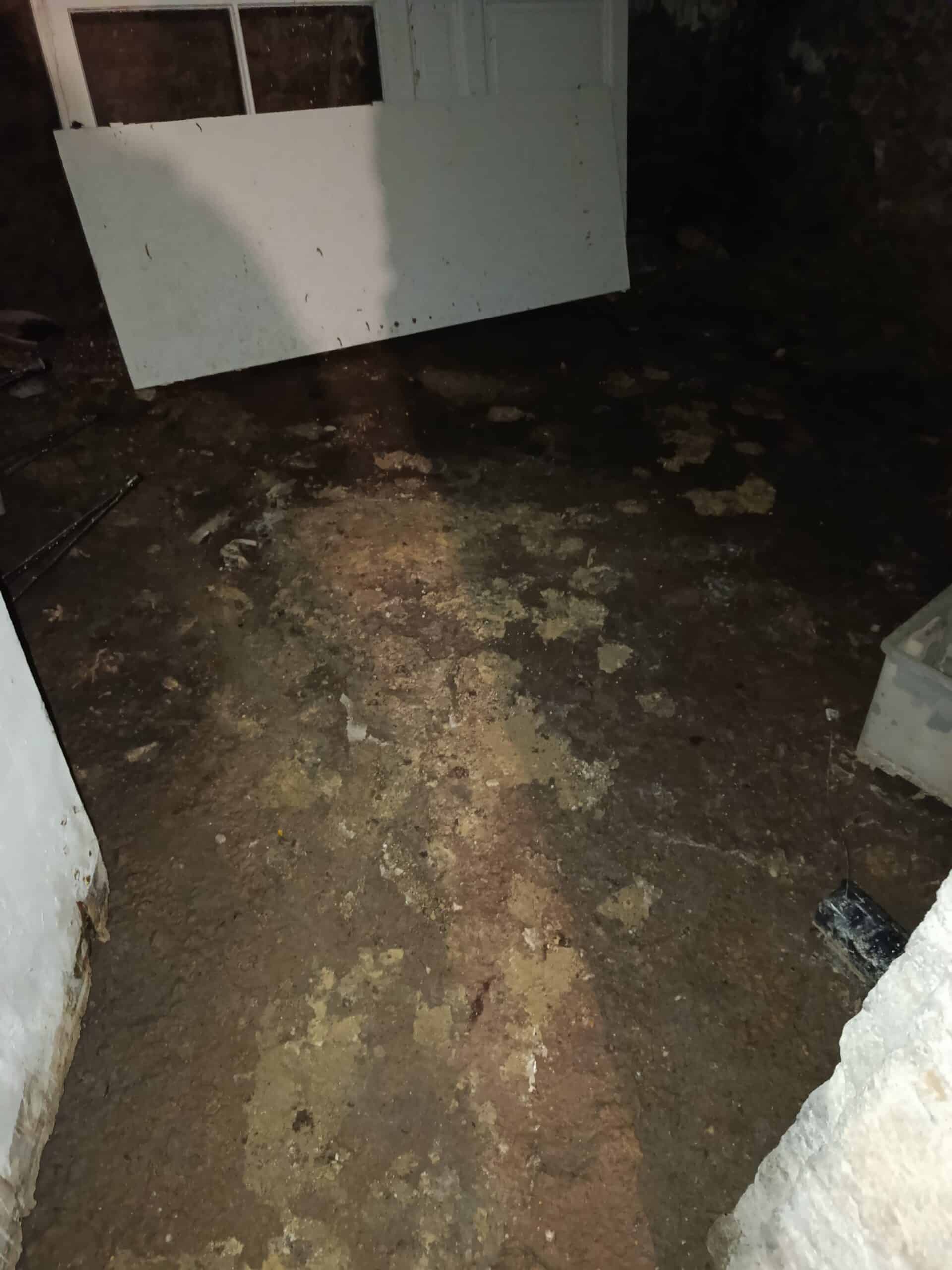 Sol boueux et humide d'un sous-sol sombre, montrant des dégâts des eaux, avec une planche blanche inclinée et des résidus jaunes.