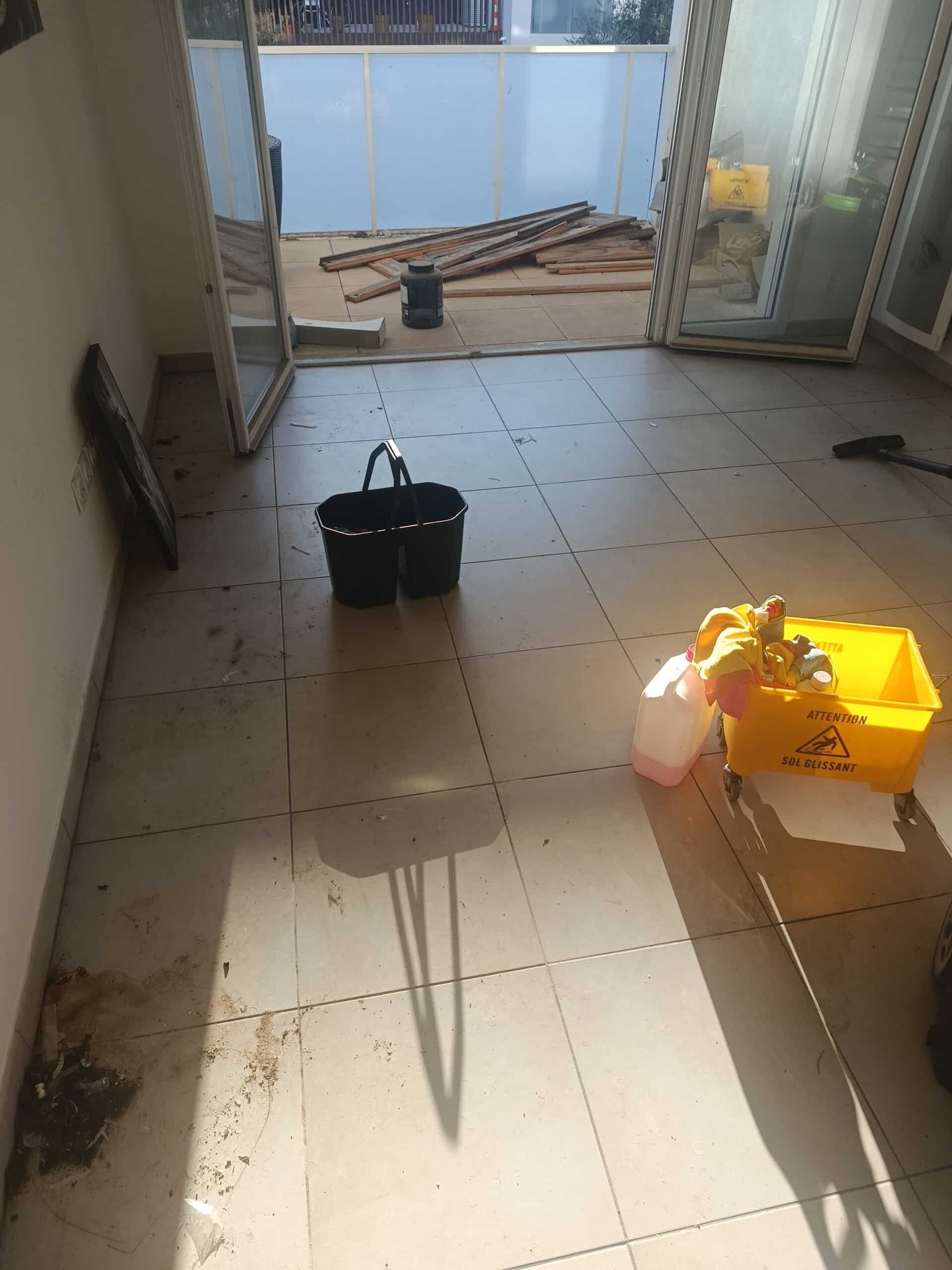 Seaux de ménage et chariot Attention Sol Glissant dans une pièce sale avec balcon encombré de bois.