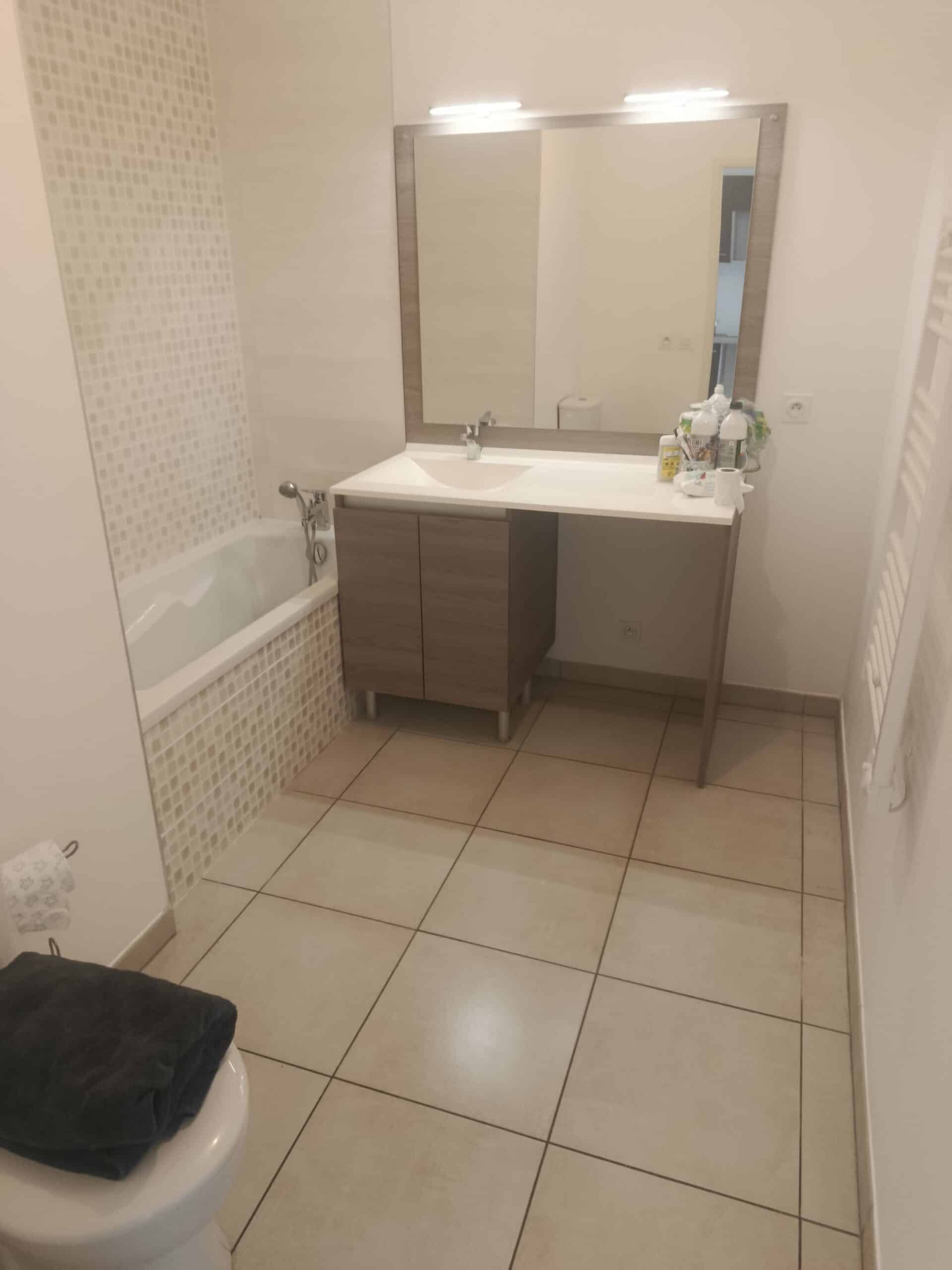 Salle de bain beige avec baignoire en mosaïque, meuble vasque en bois et grand miroir mural éclairé.