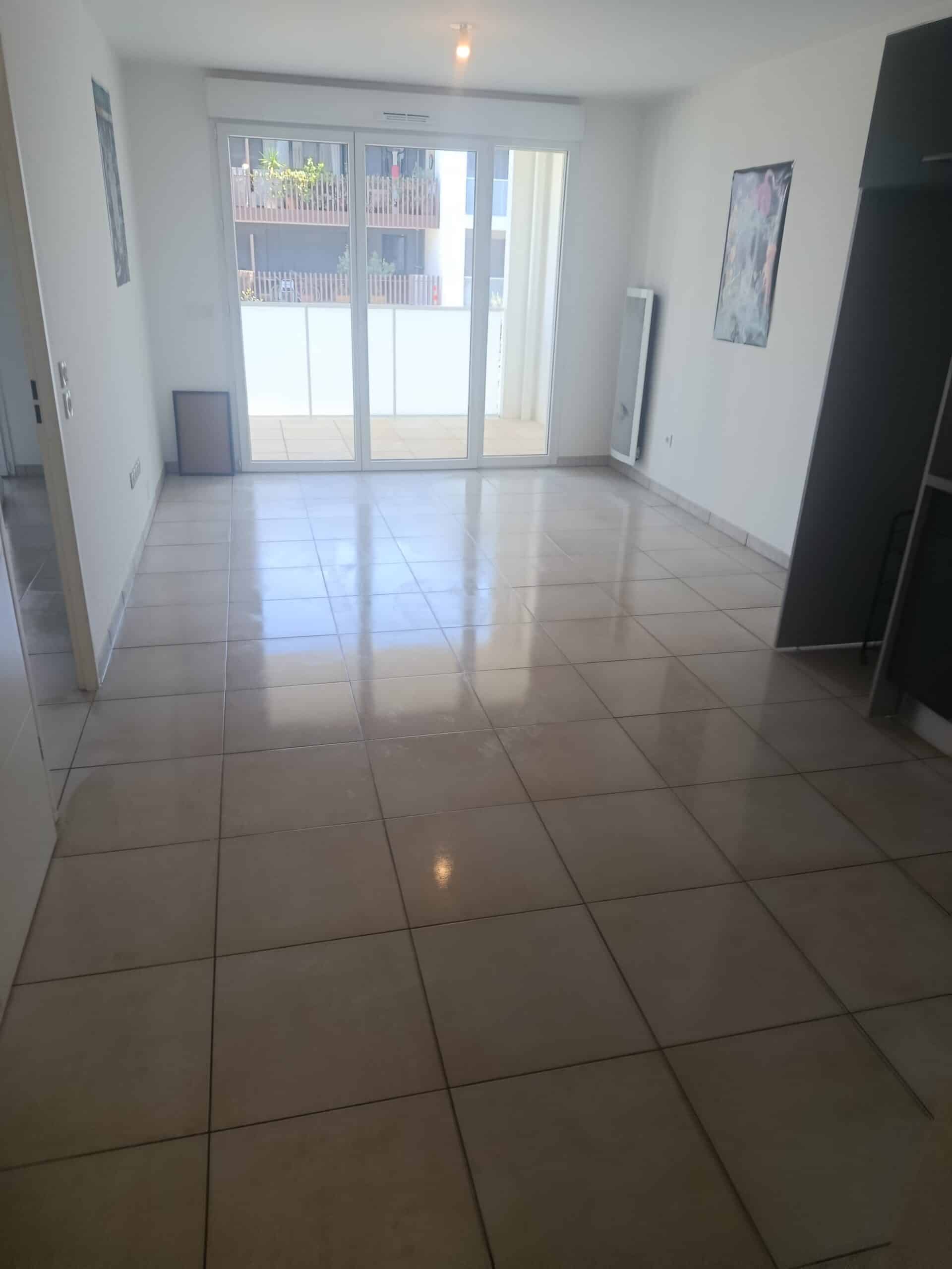 Salon vide et lumineux avec carrelage brillant, grande baie vitrée et accès à un balcon extérieur.