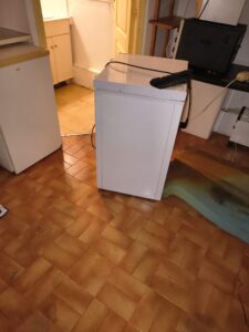Congélateur blanc compact posé sur un sol en carrelage brun dans une cuisine avec divers appareils.