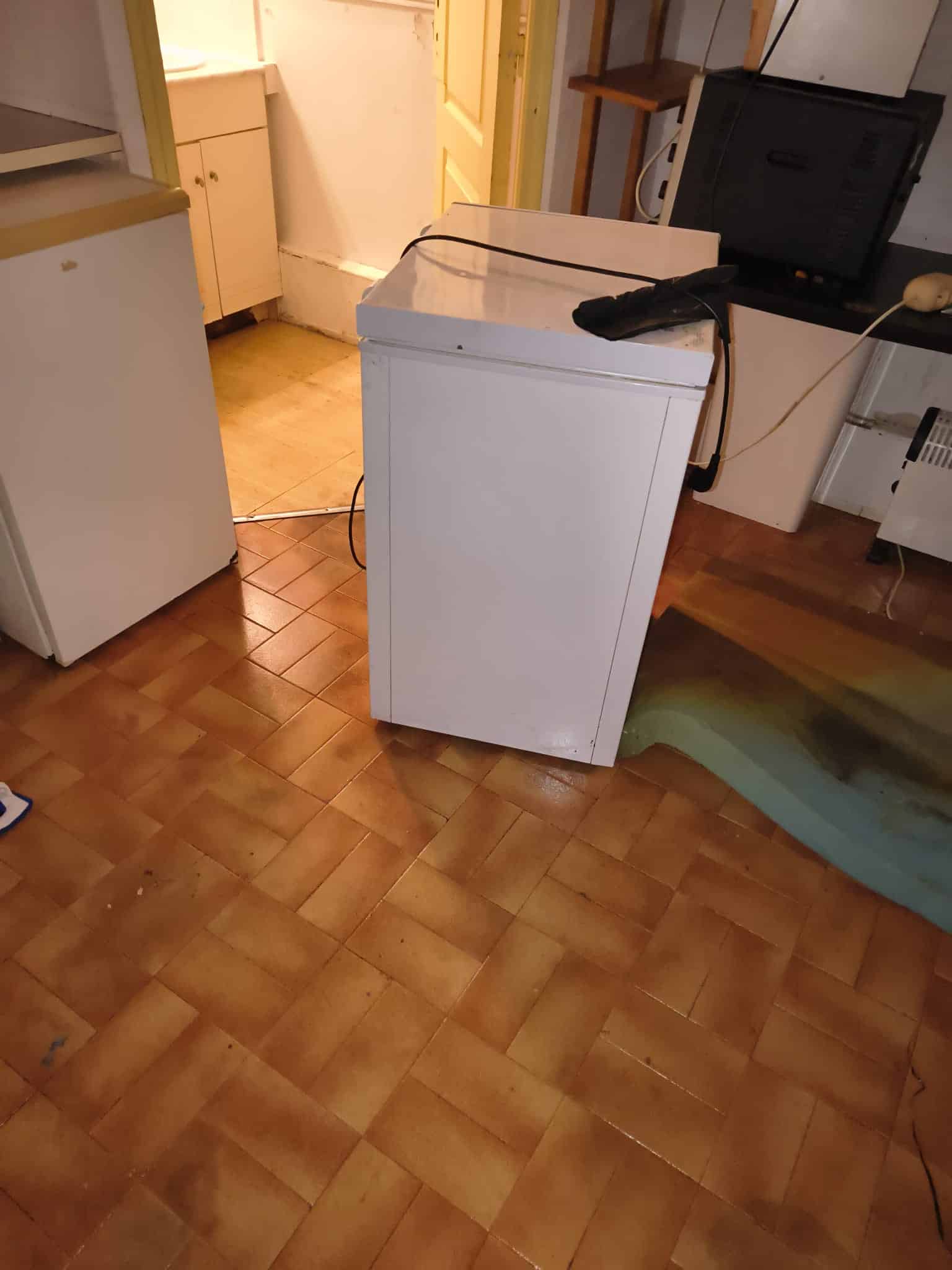 Congélateur blanc compact posé sur un sol en carrelage brun dans une cuisine avec divers appareils.