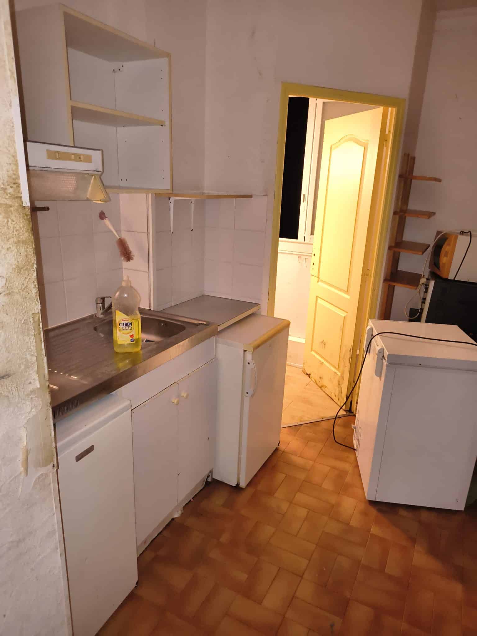 Petite cuisine exiguë avec évier inox, savon CITRON, frigo et porte jaune ouverte.