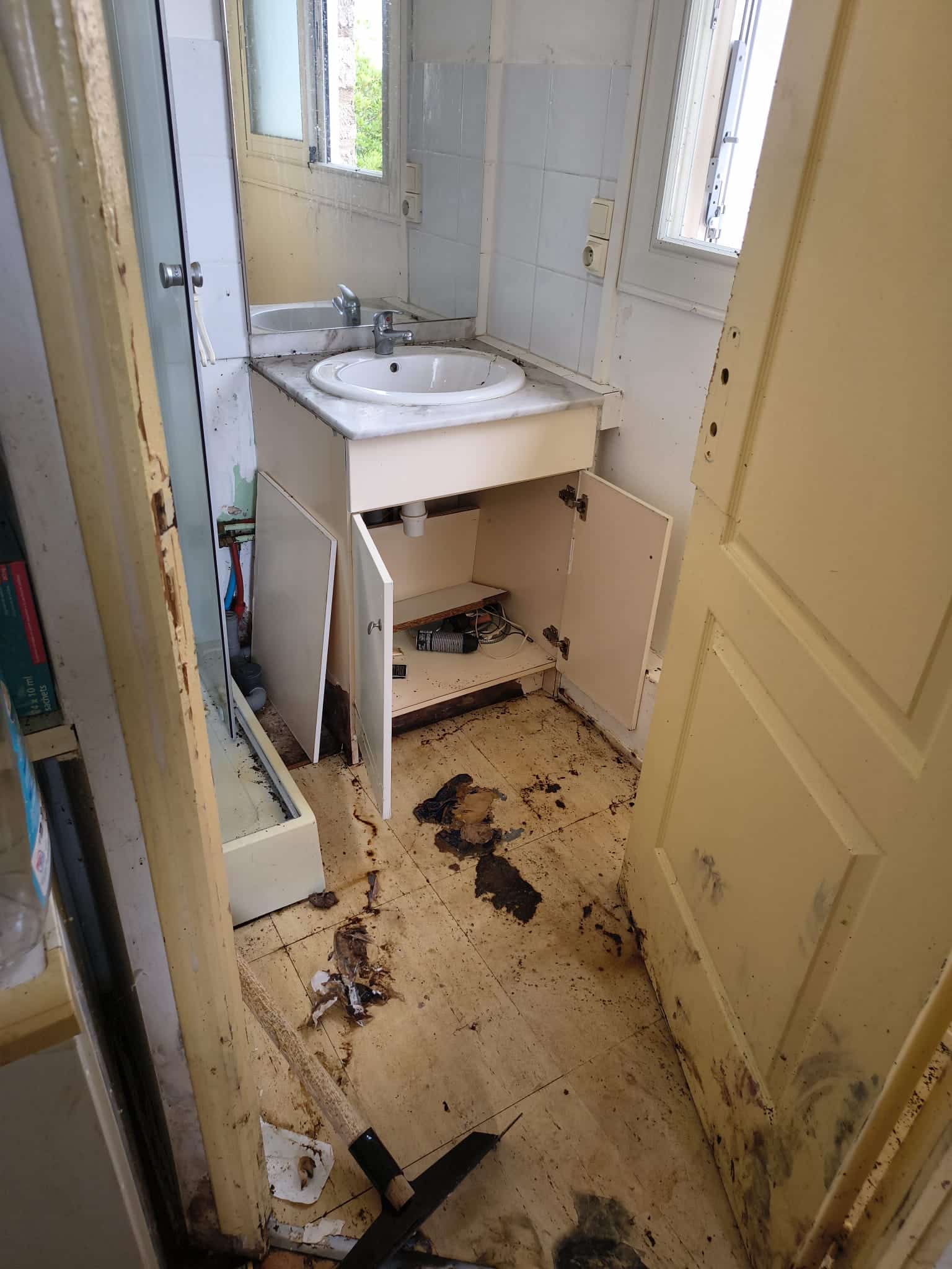 Salle de bain insalubre, sol taché, meuble ouvert et débris. État de délabrement avant travaux de rénovation.