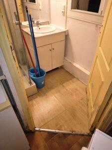 Salle de bain dégradée avec meuble vasque blanc, seau de ménage bleu et carrelage taché à rénover.