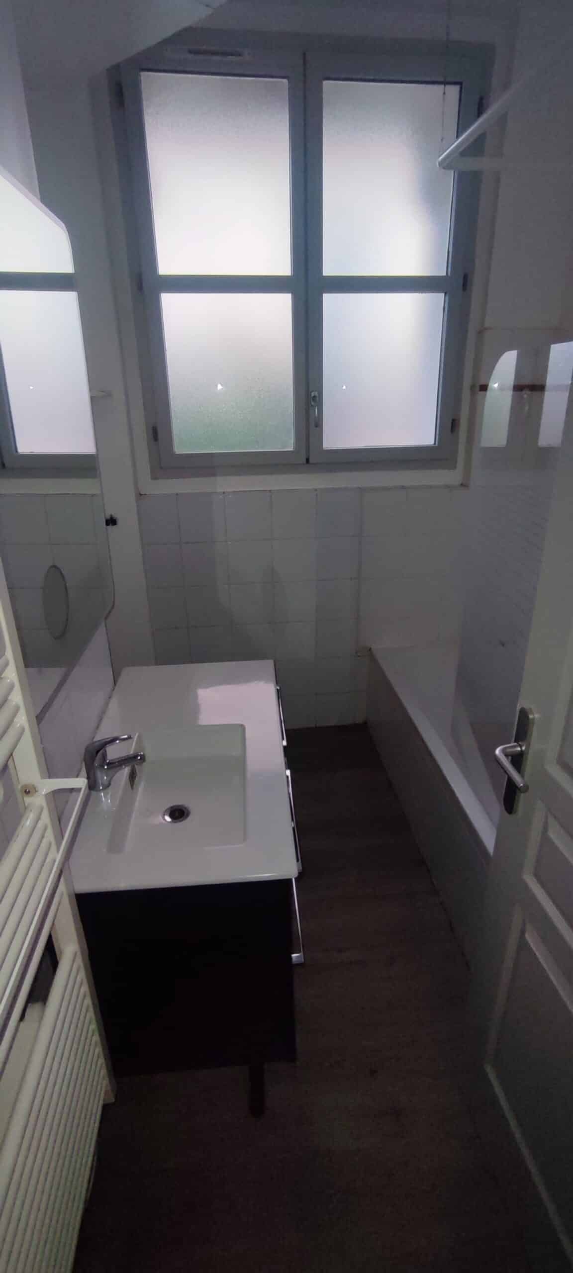 Salle de bain moderne avec meuble vasque noir, baignoire blanche, fenêtre dépolie et sol effet bois sombre.