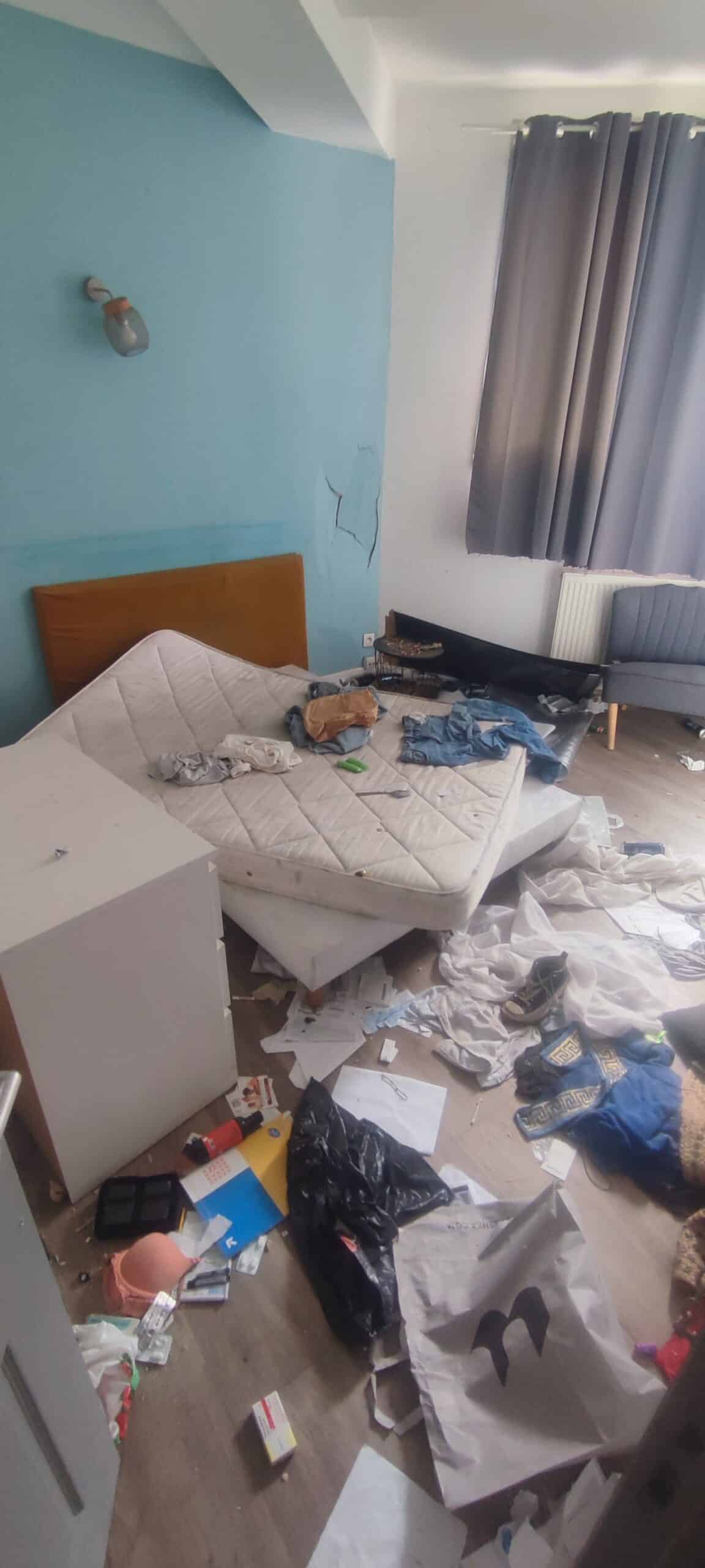 Chambre insalubre : matelas de travers, vêtements et détritus éparpillés sur le sol près d'un mur bleu fissuré.