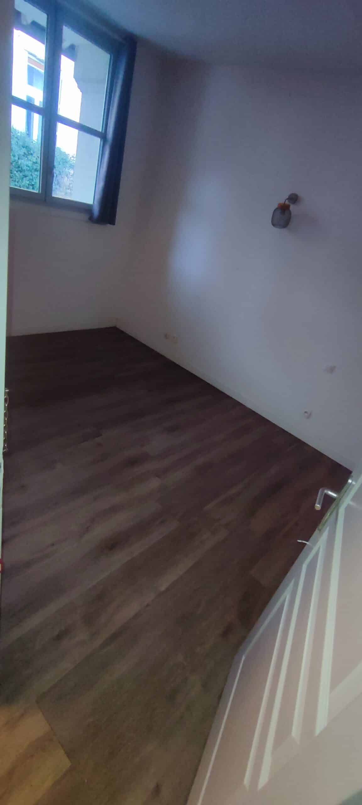 Chambre vide avec parquet en bois sombre, murs blancs, fenêtre lumineuse et petite applique murale.