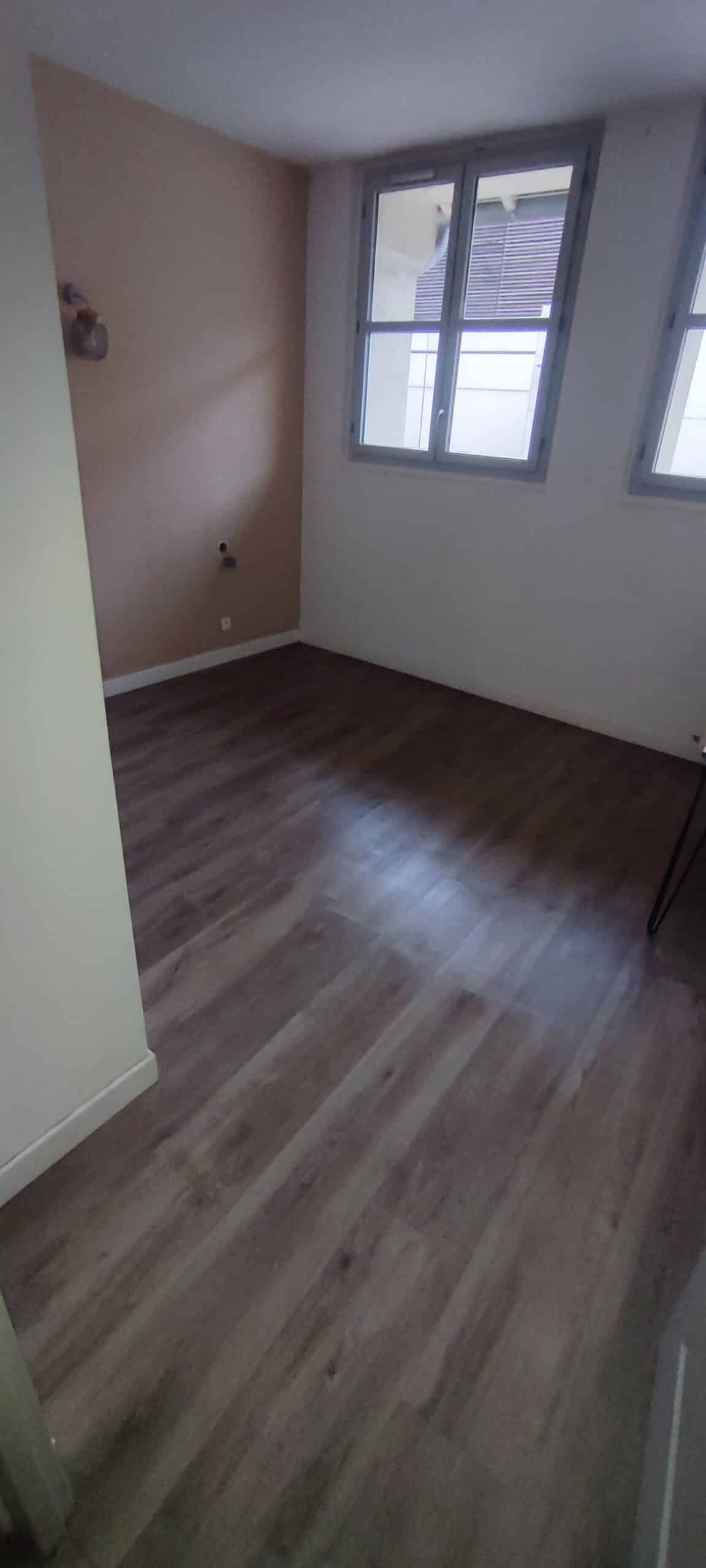 Chambre vide avec sol stratifié effet bois, un mur terracotta et deux fenêtres à cadres gris.