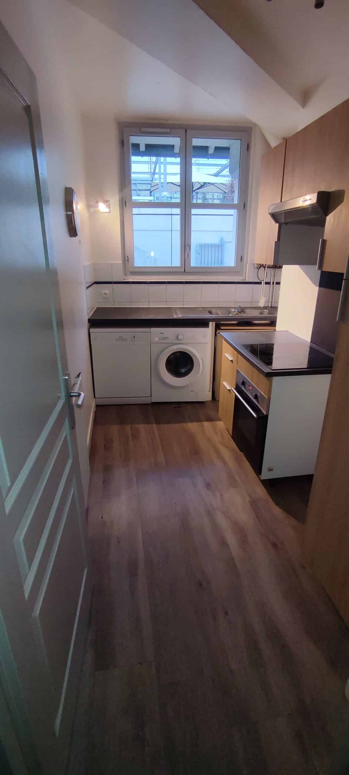 Petite cuisine moderne équipée avec lave-linge, lave-vaisselle, four et fenêtre sur sol en parquet bois.