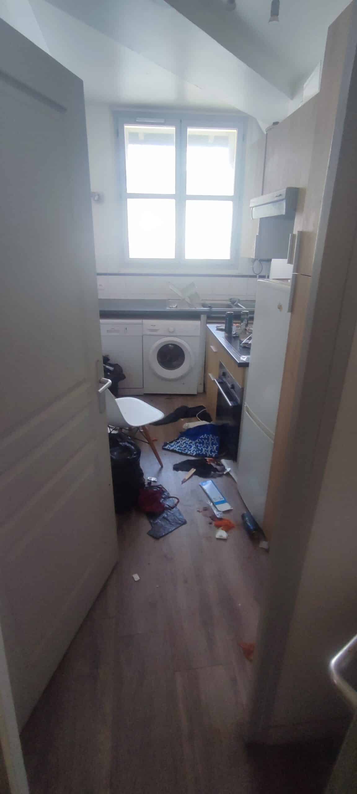Cuisine en désordre avec déchets au sol, lave-linge et frigo blanc, vue à travers une porte ouverte.