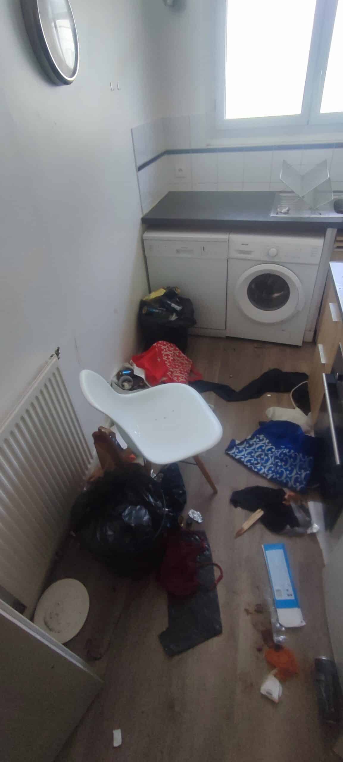 Cuisine en désordre avec sacs poubelle, linge au sol, chaise blanche et lave-linge sous un plan de travail.
