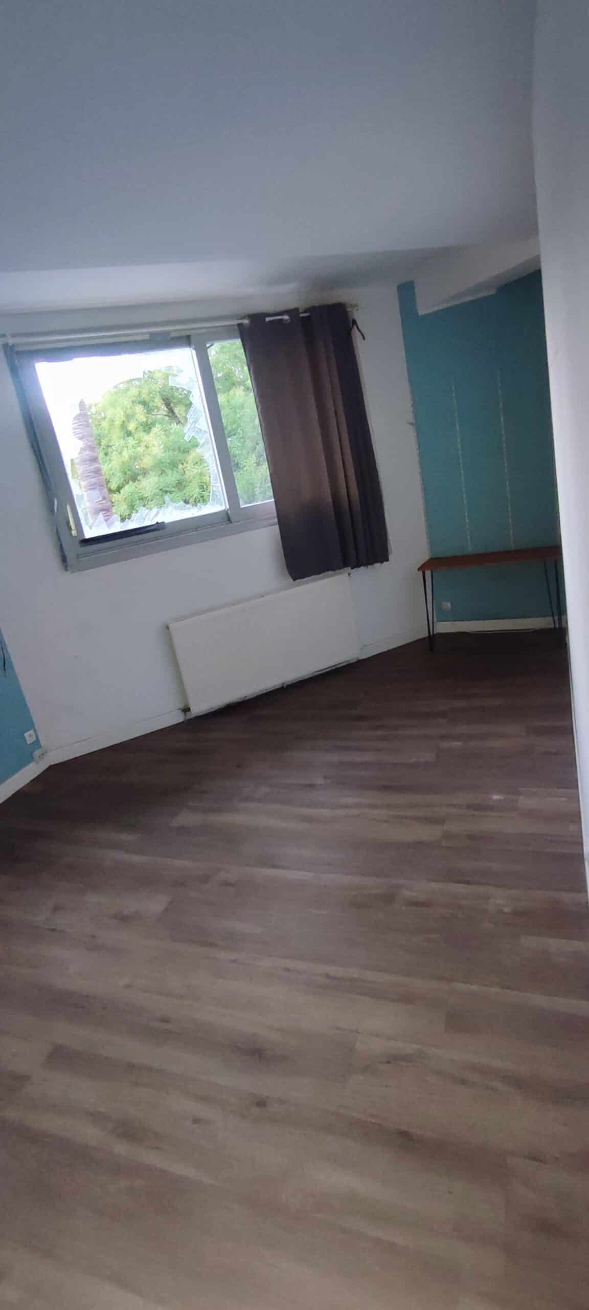 Chambre avec sol en bois, fenêtre brisée, rideaux marron et mur bleu. Pièce vide nécessitant des rénovations.