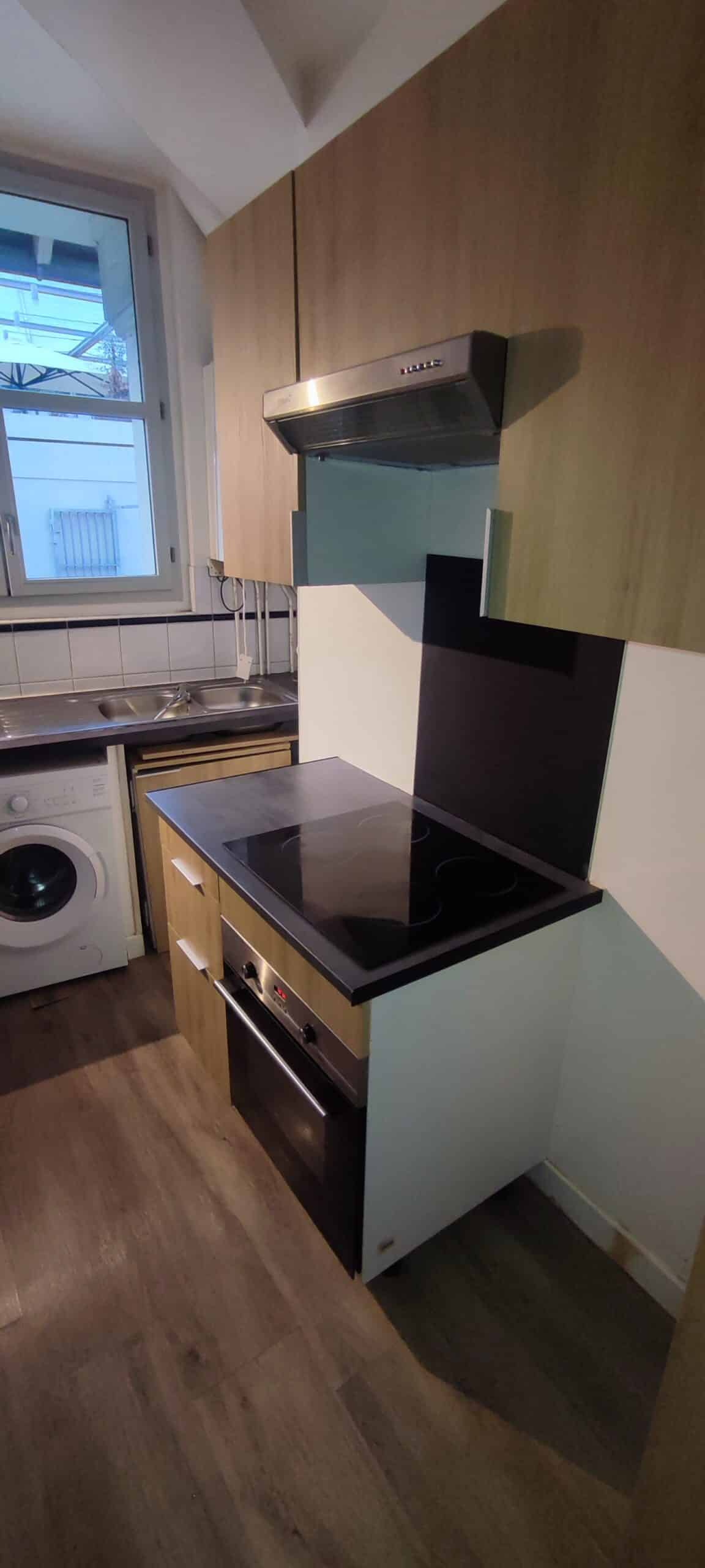 Petite cuisine moderne avec plaques à induction, four noir, lave-linge blanc et placards en bois clair.