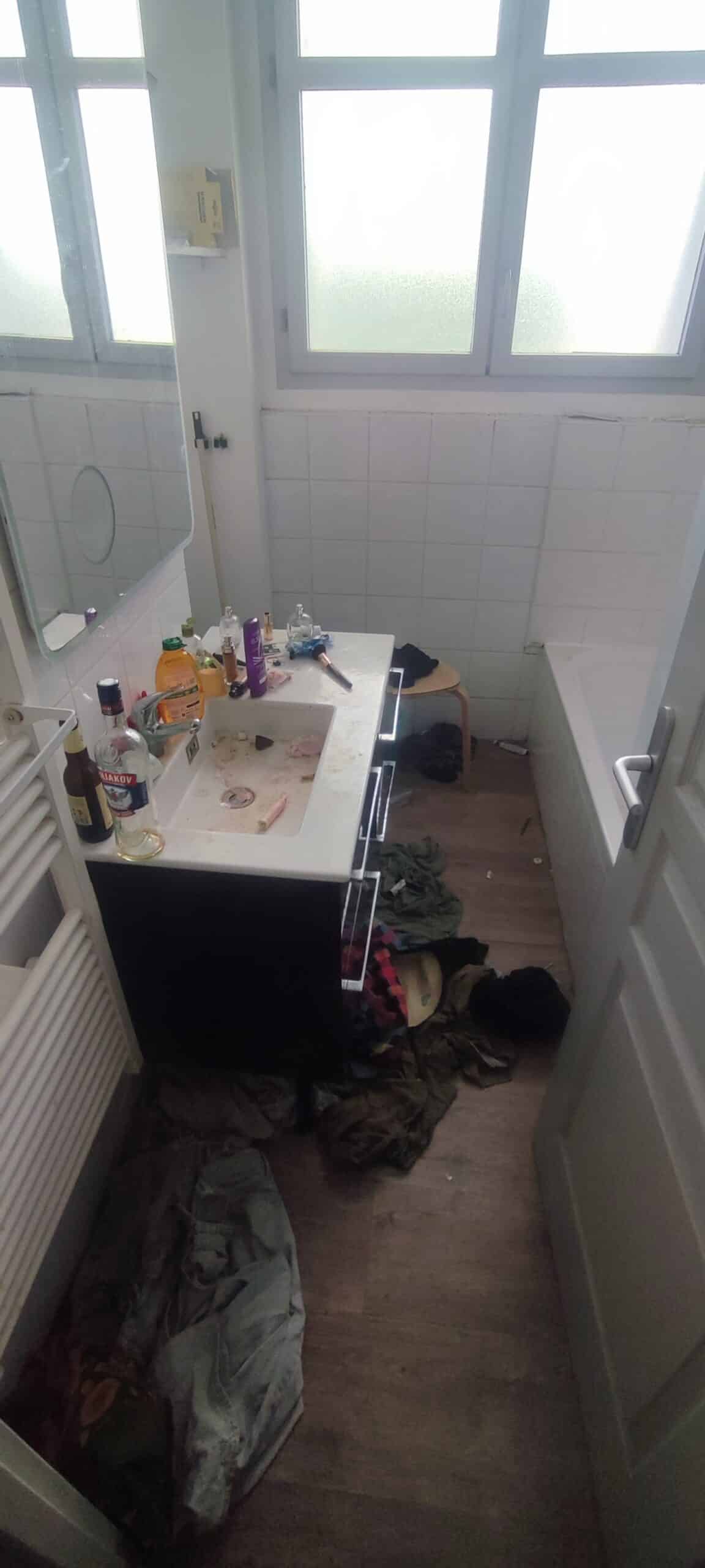 Salle de bain en désordre avec vêtements au sol, lavabo encombré de bouteilles d'alcool et baignoire blanche.